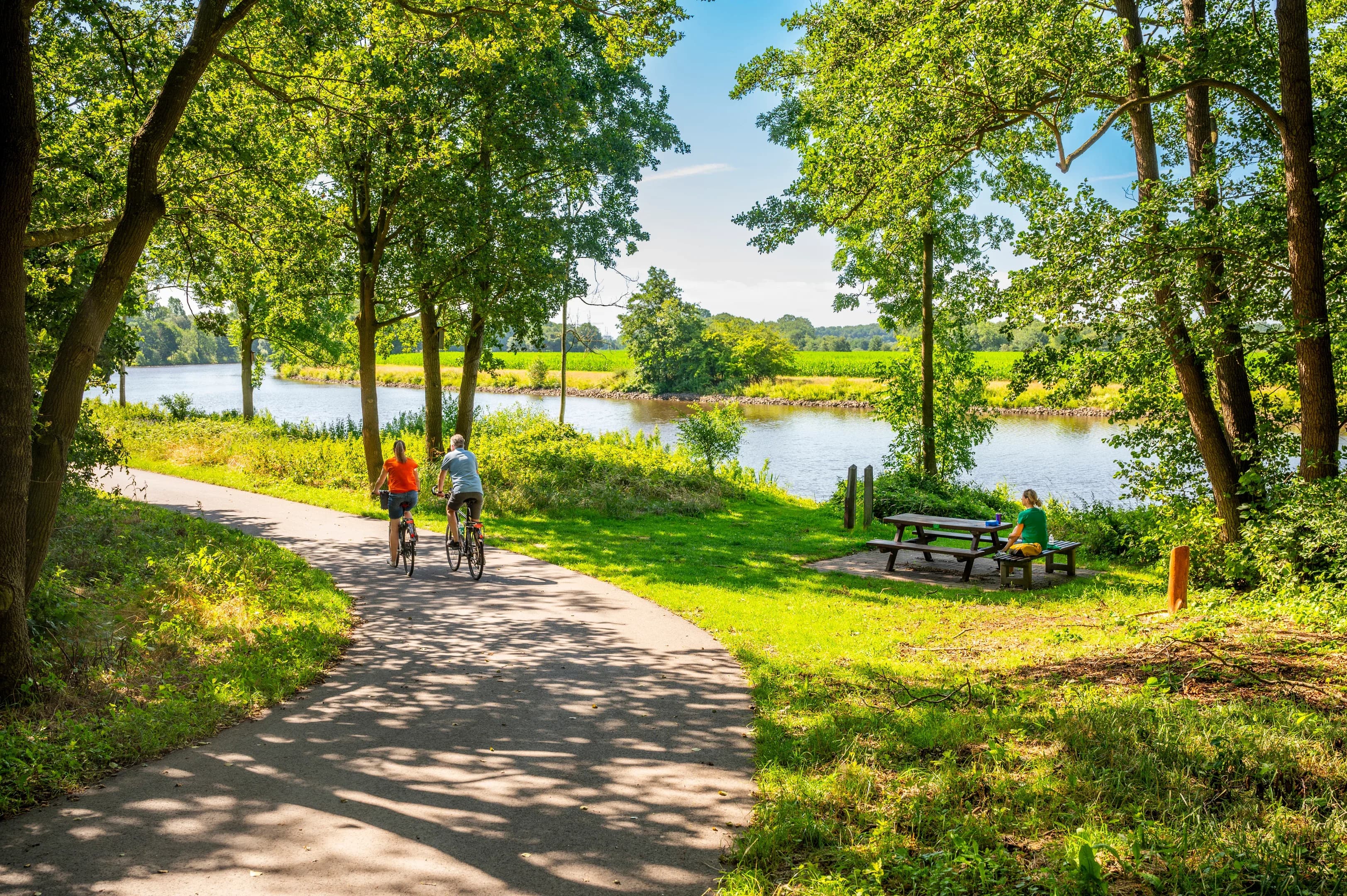 Zwei Radfahrende auf einem schattigen Weg am Kanal, daneben eine Picknickbank