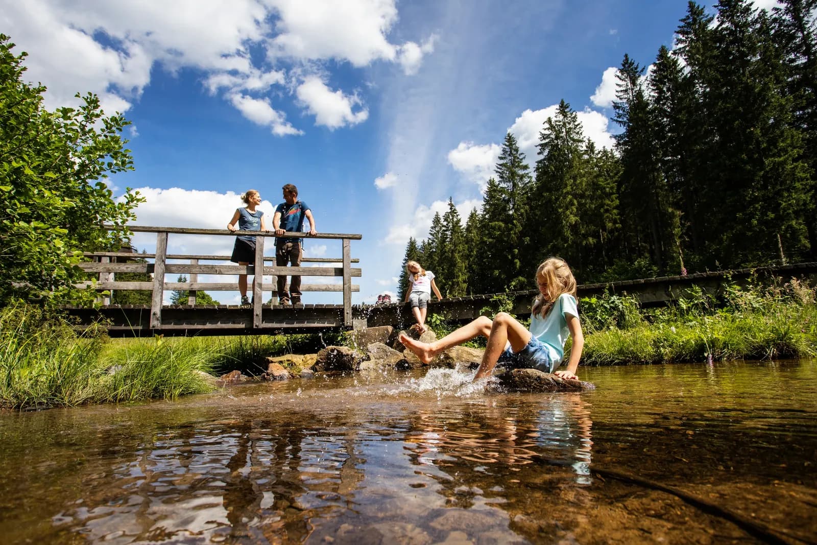 Kinder spielen im flachen Wasser eines Baches, Erwachsene stehen auf der Brücke darüber